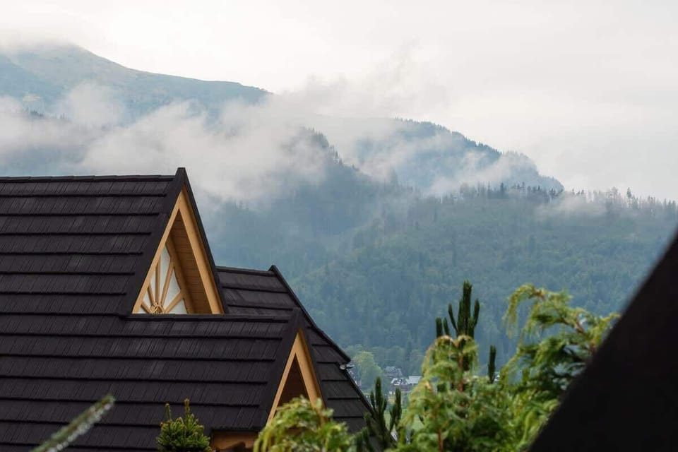 A mountain view framed by rooftops and treetops, with mist rolling over the peaks.

