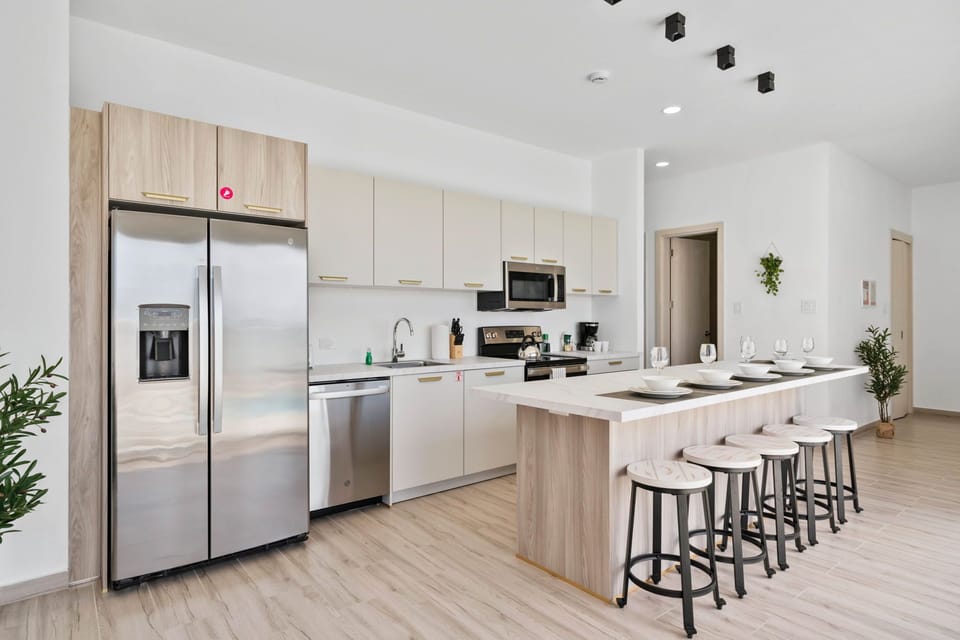- Modern kitchen featuring stainless steel appliances and minimalist cabinetry - Spacious island with stools set for five, perfect for casual dining or entertaining - Clean, neutral tones create a bright and airy atmosphere, enhanced by natural light