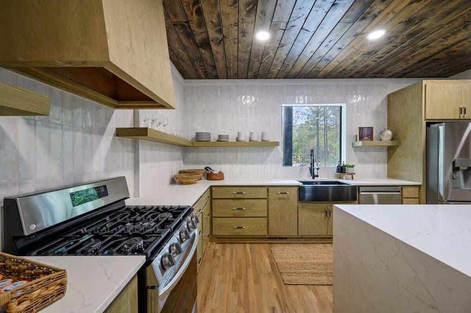 Stylish kitchen with wood cabinetry, stainless steel appliances, farmhouse sink, open shelving, and natural light through a large window with forest views.