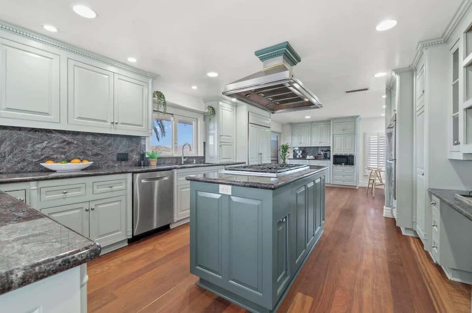 This kitchen blends modern elegance with functionality—top-of-the-line built-in appliances, sleek counters, and white cabinetry. A pop of color on the island brightens the space, while a window by the sink brings in stunning views.