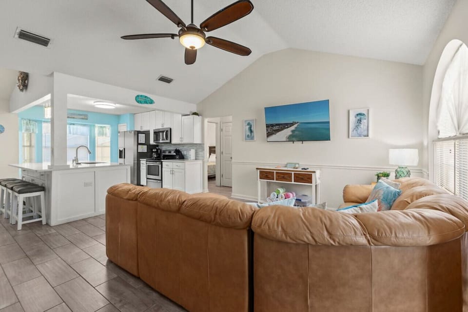 A spacious living room with tall ceilings, a comfortable leather sectional, and a beach theme, filled with natural light and an open, airy feel.