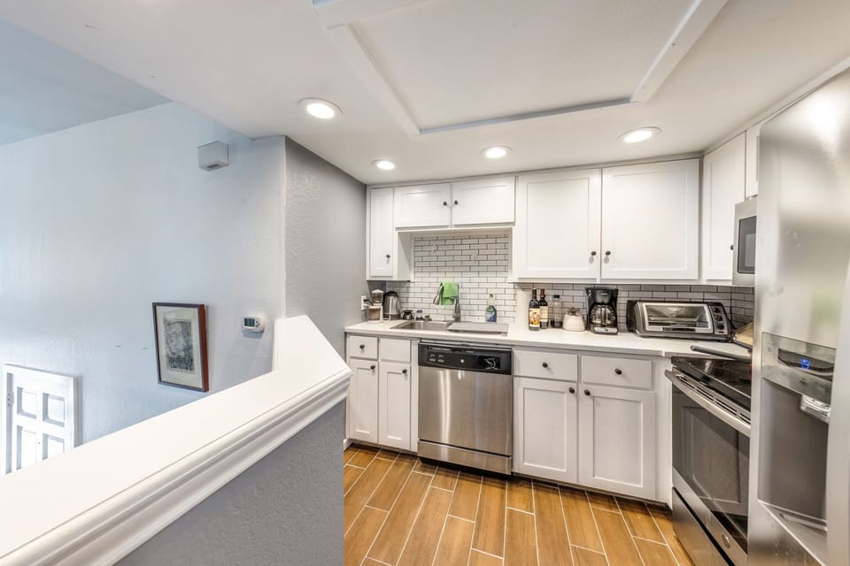 A modern kitchen with white cabinets, stainless steel appliances, a tiled backsplash, and wooden flooring, viewed from a slightly elevated perspective.