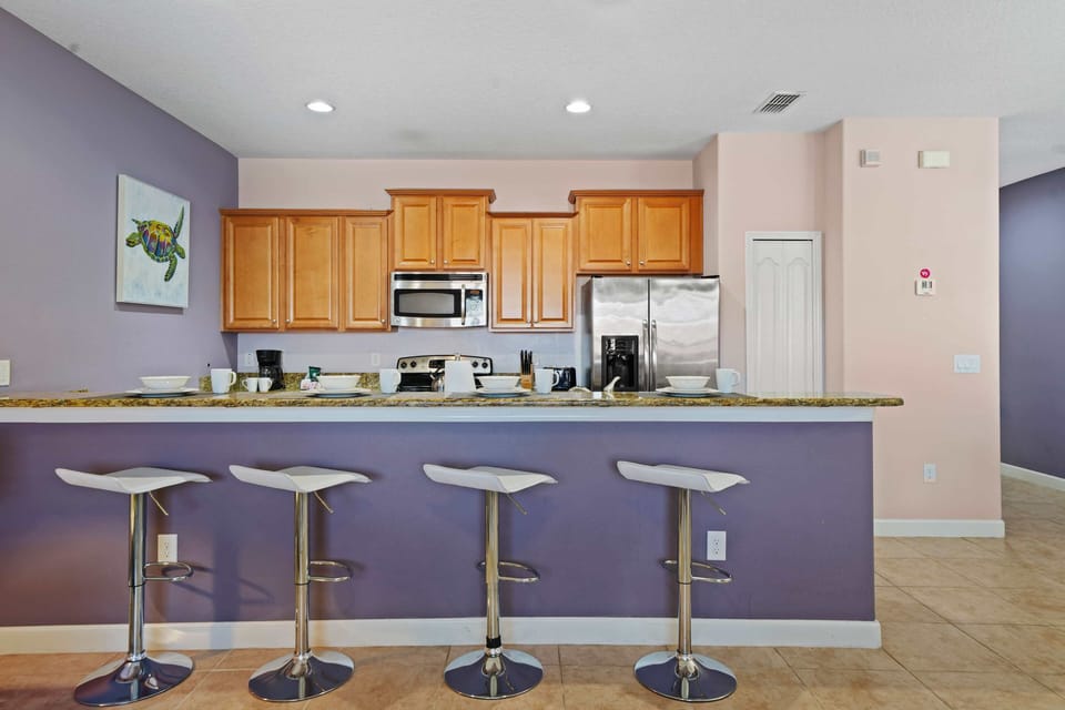 - Spacious kitchen with warm wood cabinetry, granite countertops, and stainless steel appliances - Breakfast bar with four modern white stools against a soft purple accent wall - Coastal art adds a playful touch to the stylish and functional space