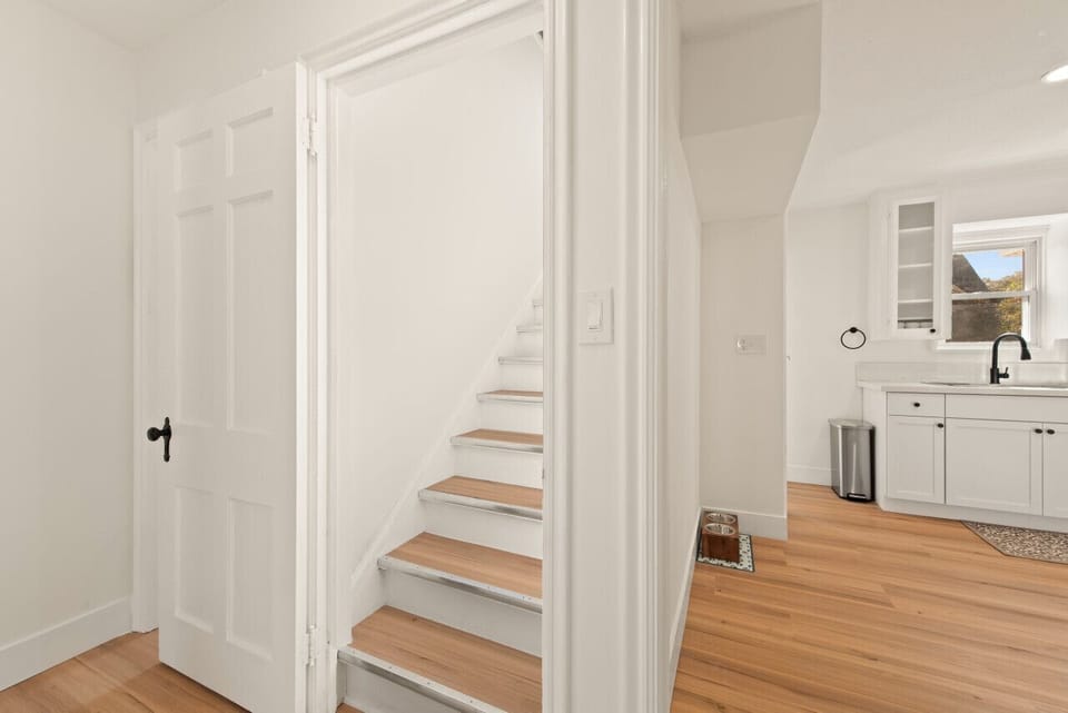- Hallway view with stairs leading to upper level bedroom retreat   - White walls and wood flooring keep spaces bright and seamless   - Kitchen entry nearby creates easy flow through the home