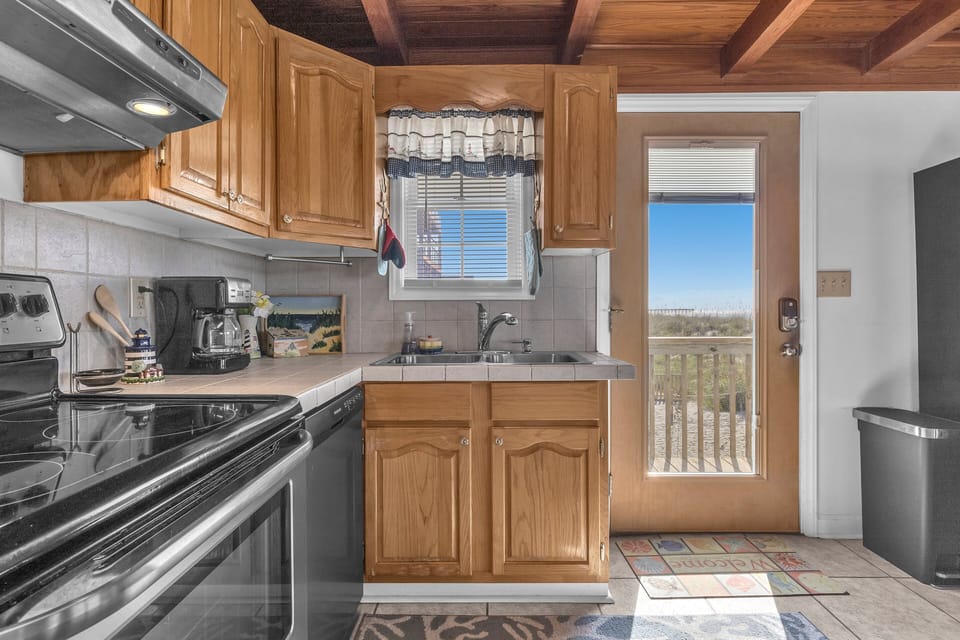 Cozy kitchen with wood cabinets, stainless appliances, tile counters, double sink, microwave, coffee maker, wood ceiling beams, coral rug, and ocean-view door.