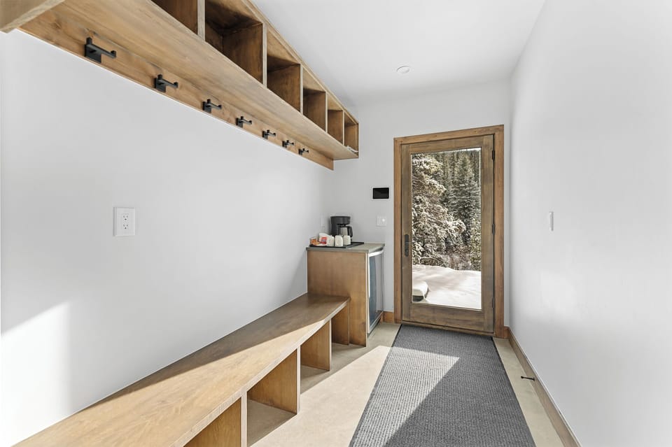 Your private mudroom features warm wood storage and bench seating, with coffee station and glass door to snowy outdoor retreat.