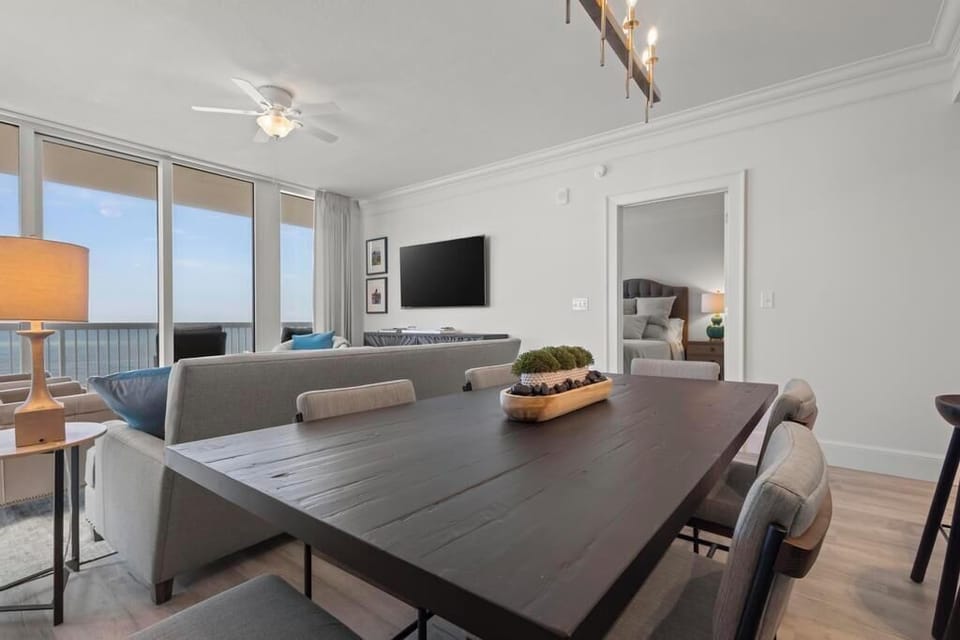 The dining area features a large dining table, a cozy sofa, and a TV, with a stunning beach view through expansive windows