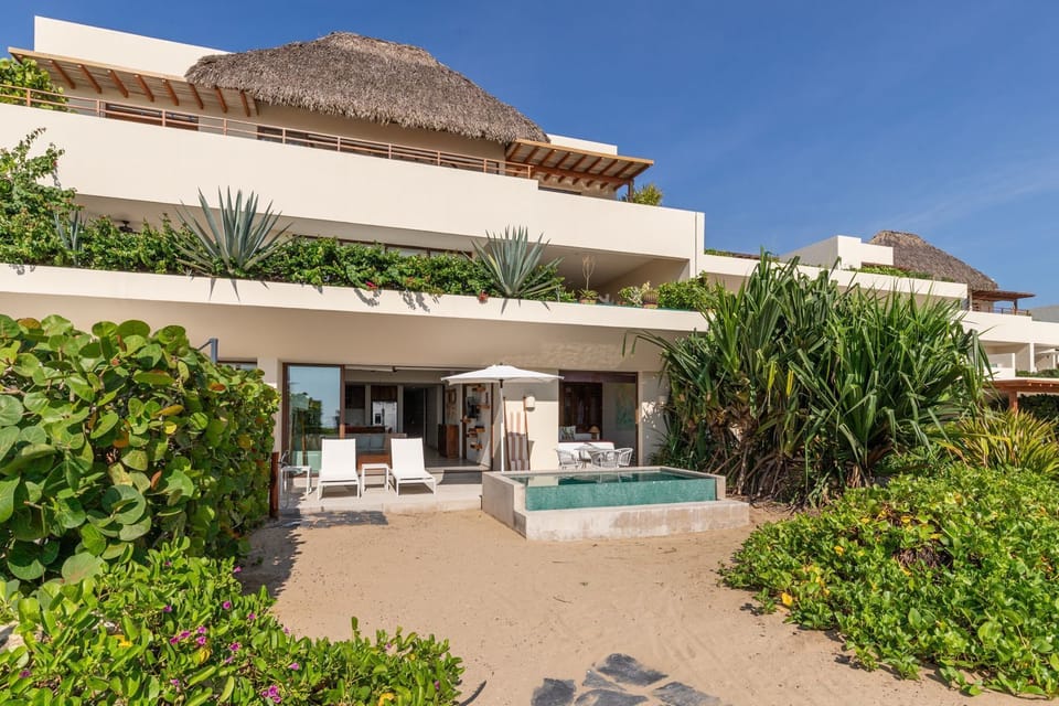 A stunning beachfront villa featuring a private pool, lush tropical landscaping, and shaded outdoor lounge areas.