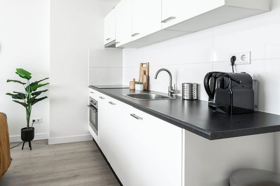 Modern kitchen featuring sleek white cabinetry, black countertops, and stainless steel appliances. Equipped with an oven, sink, and a coffee machine, this space offers functionality and style. A potted plant adds a fresh touch, making it perfect for travelers seeking a fully equipped, contemporary kitchen.