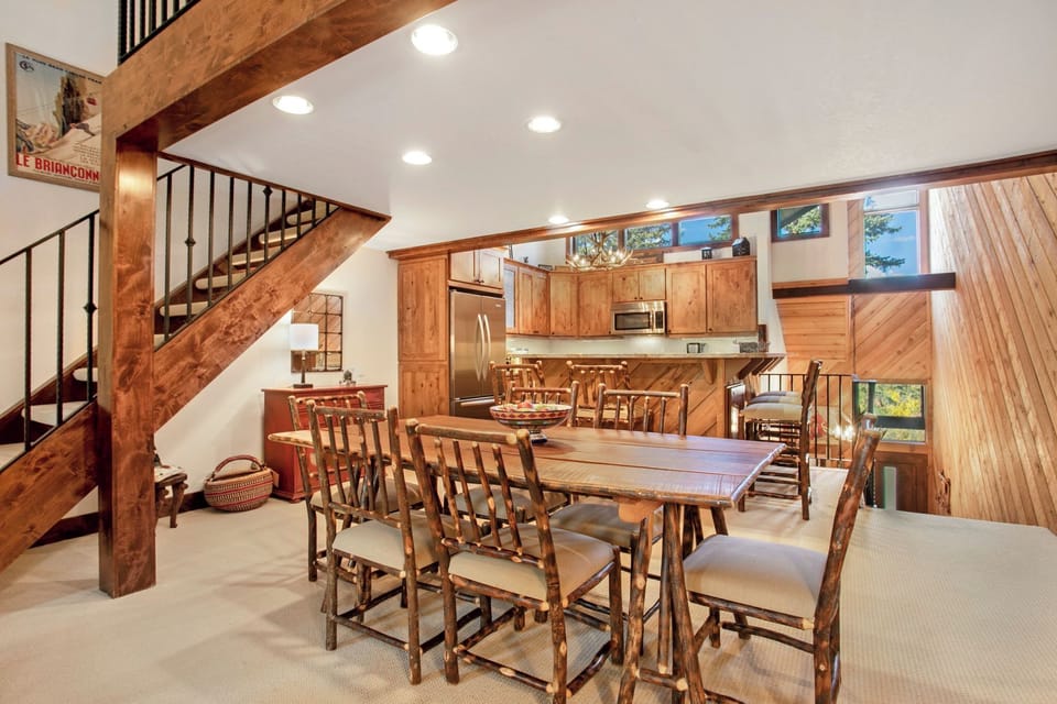 A rustic dining area with a wooden table and chairs, adjacent to a kitchen with wooden cabinets and stainless steel appliances. A staircase leads to an upper level. Natural light enters through high windows.