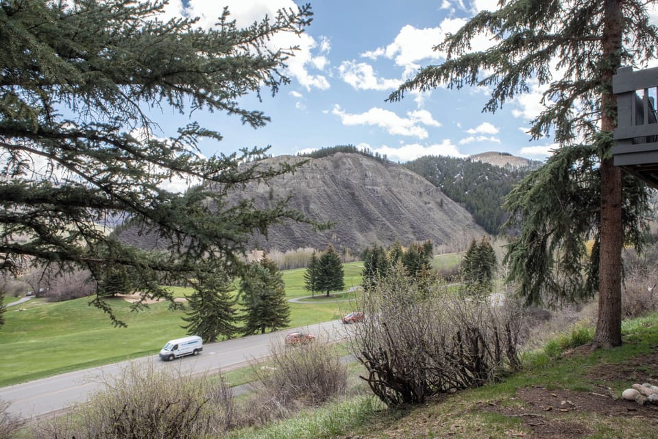 A road runs through a lush valley lined with evergreen trees, with a large rocky hill and a cloudy blue sky in the background. Vehicles are visible on the road.