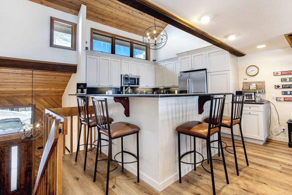 A modern white kitchen with high wooden ceilings, bar seating for four, stainless steel appliances, and large windows providing natural light. The space is adjacent to a wooden staircase.