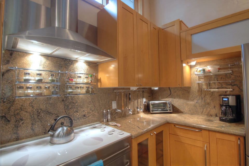 Modern kitchen with stainless steel appliances, wooden cabinets, granite countertops, a stove with a kettle, a toaster oven, and a coffee maker under bright lighting.