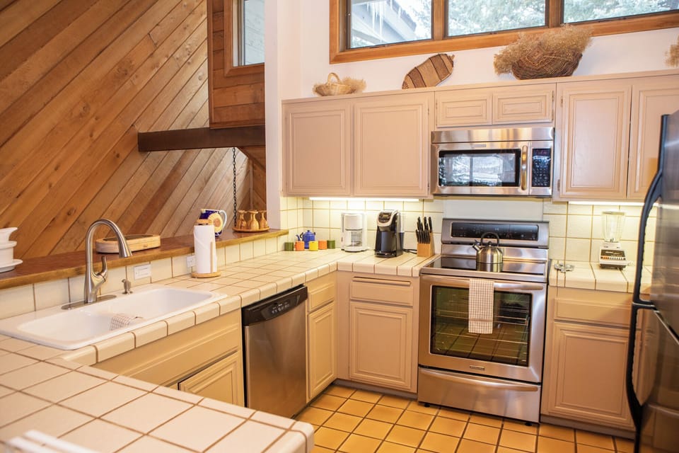 A modern kitchen with beige cabinetry, stainless steel appliances, a tiled countertop, and a diagonal wood accent wall. Various utensils and decor items are neatly arranged on the counters.