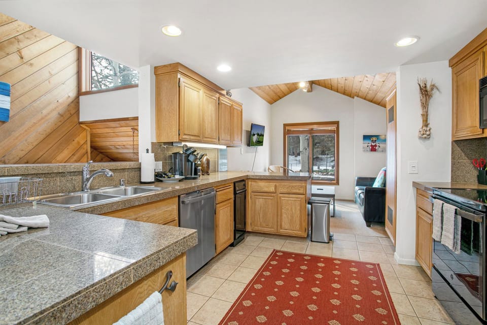 A modern kitchen with wooden cabinets and granite countertops, featuring stainless steel appliances. The kitchen opens into a living area with wooden walls and ceiling, and a red patterned rug on the floor.