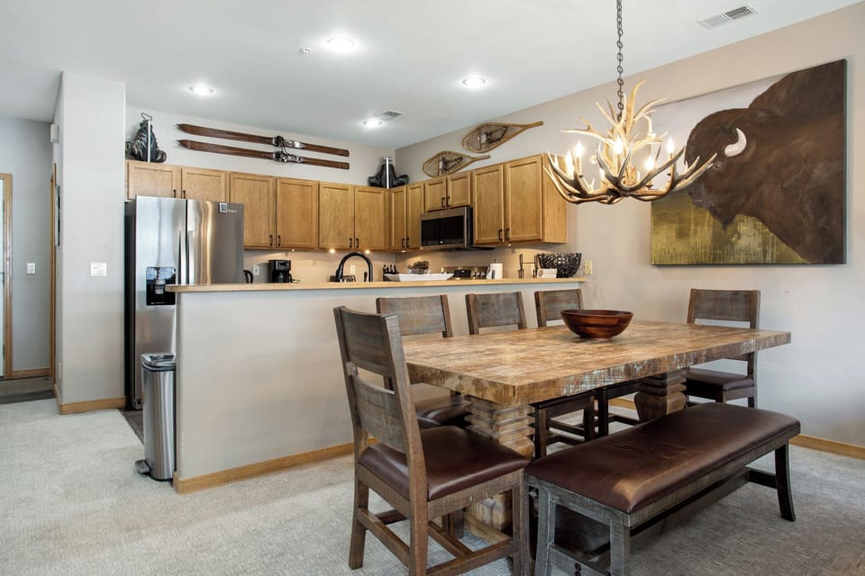 A rustic-themed kitchen and dining area featuring a wooden table, chairs, a bench, and a chandelier with antler design. The kitchen has stainless steel appliances and wooden cabinets. A bison painting decorates the wall.