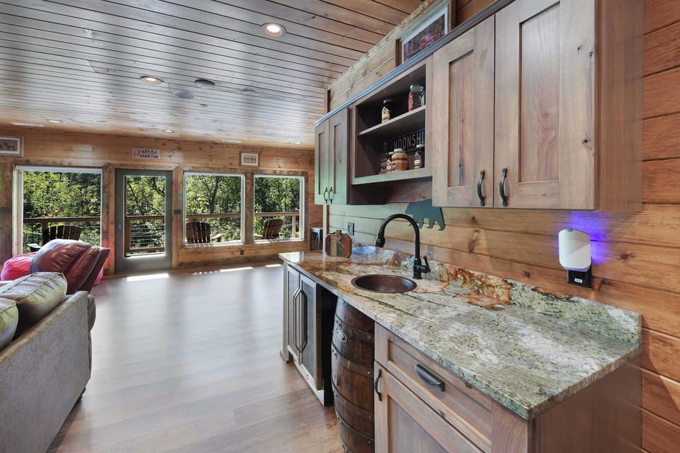 Rustic wet bar area with granite countertop and forest views through large windows.