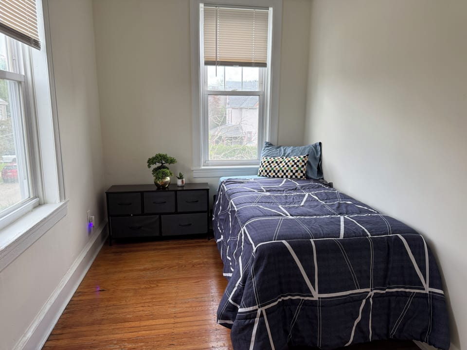 Cozy, minimalist room with a twin bed, navy geometric bedding, and a small black dresser. Bright natural light from two large windows. Simple decor includes a faux bonsai and succulent. Warm wood floors and neutral walls create a clean, airy feel.