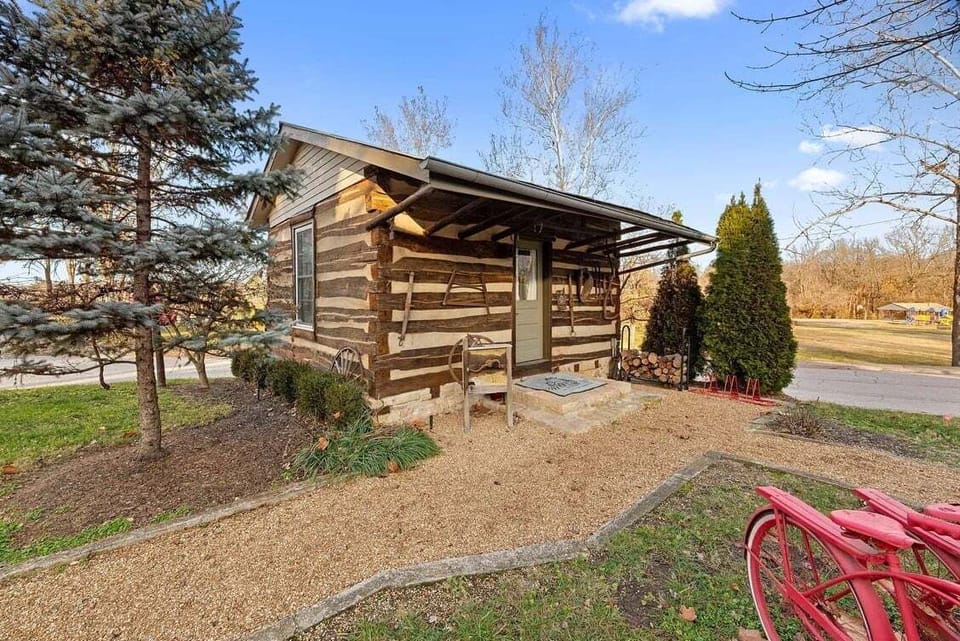 The small historic log cabin welcomes guests with a covered porch, mature trees, and a gravel walkway.