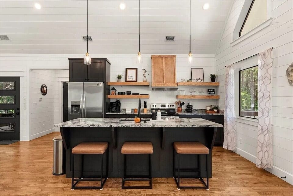 Your open kitchen has a modern farmhouse style with dark cabinets, bar stool seating, a marble-patterned island, and warm wood shelving. Pendant lighting adds elegance, while large windows and white shiplap walls create an airy, inviting atmosphere.