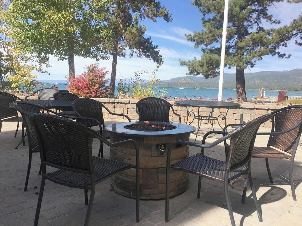 Patio seating faces the lake, beach, boats, mountains, and trees.