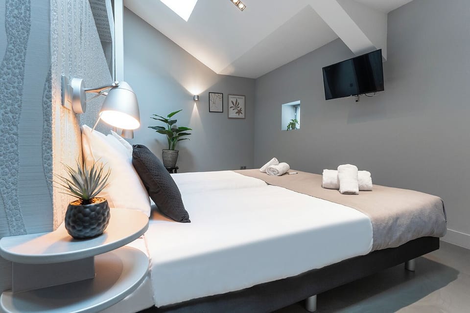 A stylish, compact bedroom under a sloped ceiling, featuring a neat bed with thoughtful touches of decor, such as a potted plant and modern bedside lighting, creating a cozy and inviting atmosphere.