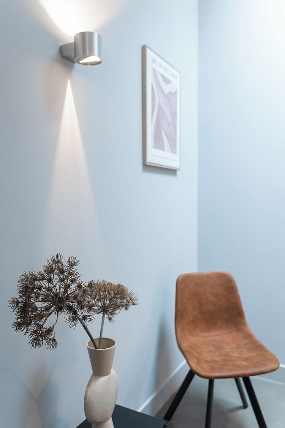 A cozy corner featuring a warm-toned chair, soft wall lighting, and a decorative vase with dried flowers, creating a tranquil and stylish ambiance.