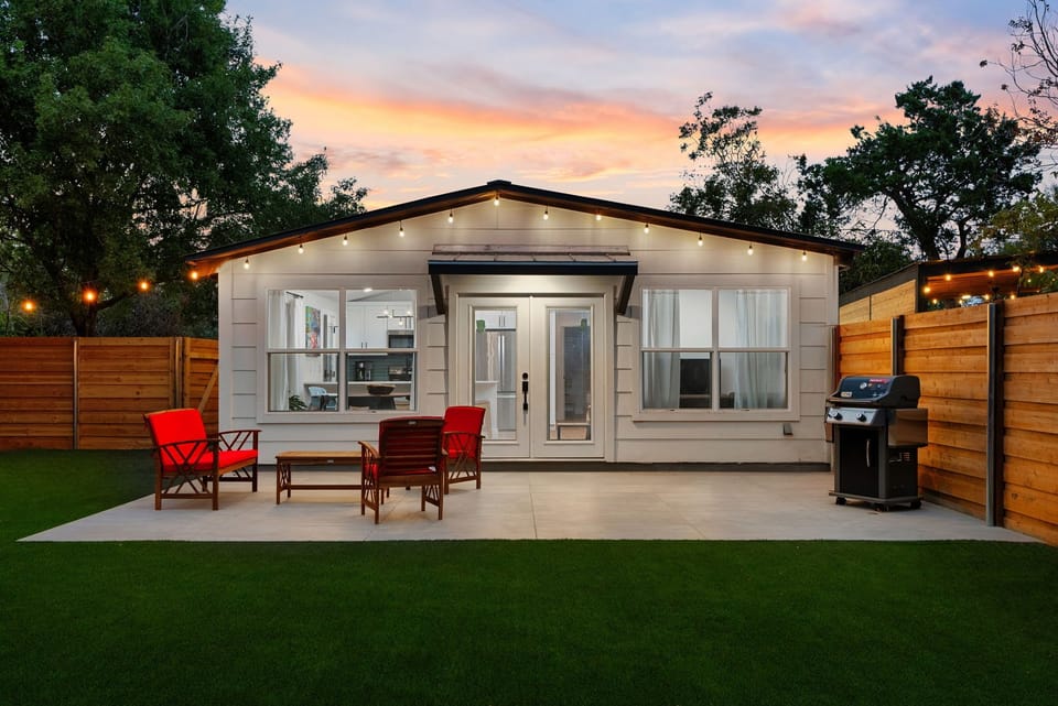This cozy, modern backyard guest house features a sleek white exterior, large windows, and a welcoming patio area with vibrant red outdoor seating.