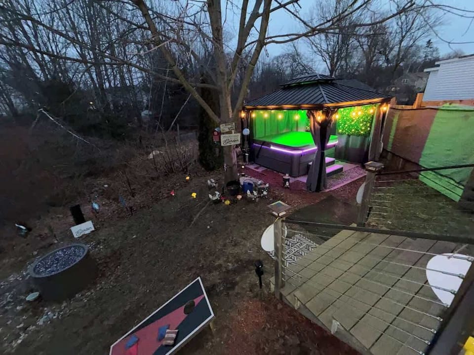 Twilight backyard overview: lit gazebo hot tub with color LEDs, fire pit, and cornhole — the whole evening lineup in one shot