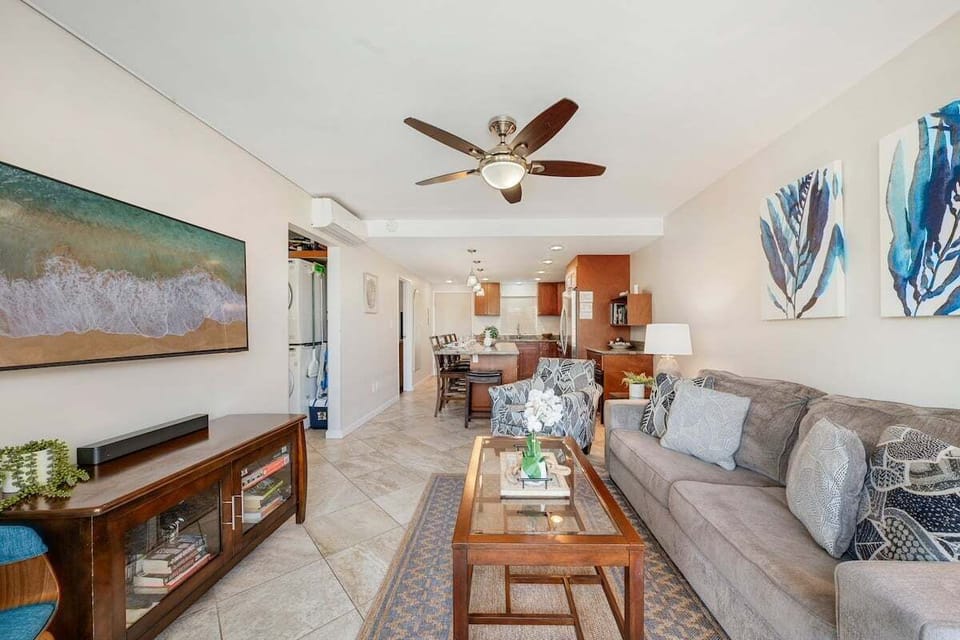 Living room with sofa, coffee table, and beach‑inspired artwork extending into dining and kitchen areas.