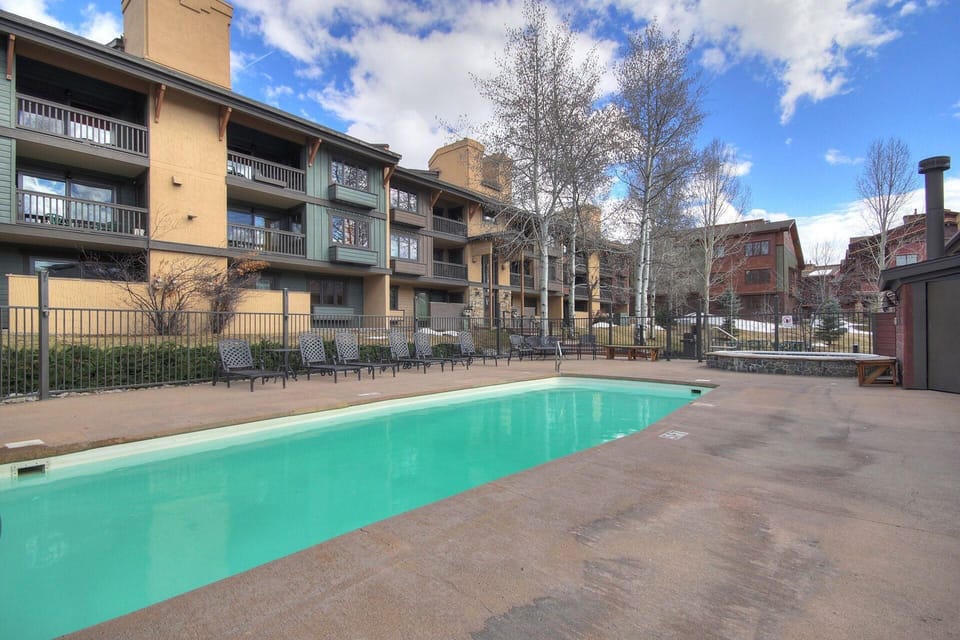 Outdoor swimming pool area with lounge chairs, surrounded by a fence and adjacent to multi-story residential buildings. Trees and a clear blue sky are visible in the background.