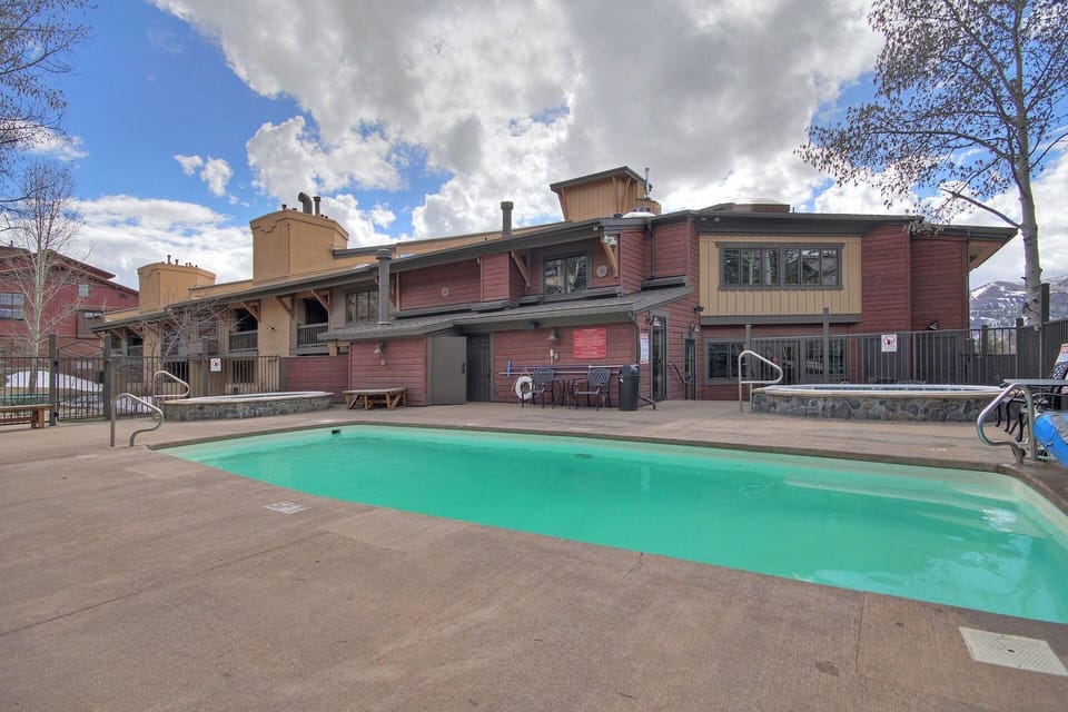 Outdoor swimming pool in front of a multi-story building with wooden and stone exterior, surrounded by trees against a partly cloudy sky.