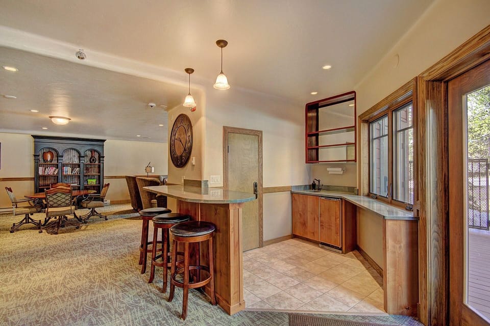 A spacious room with a small kitchen area, barstools at a counter, a dining table with chairs, bookshelves, a large wall clock, and large windows providing ample natural light.
