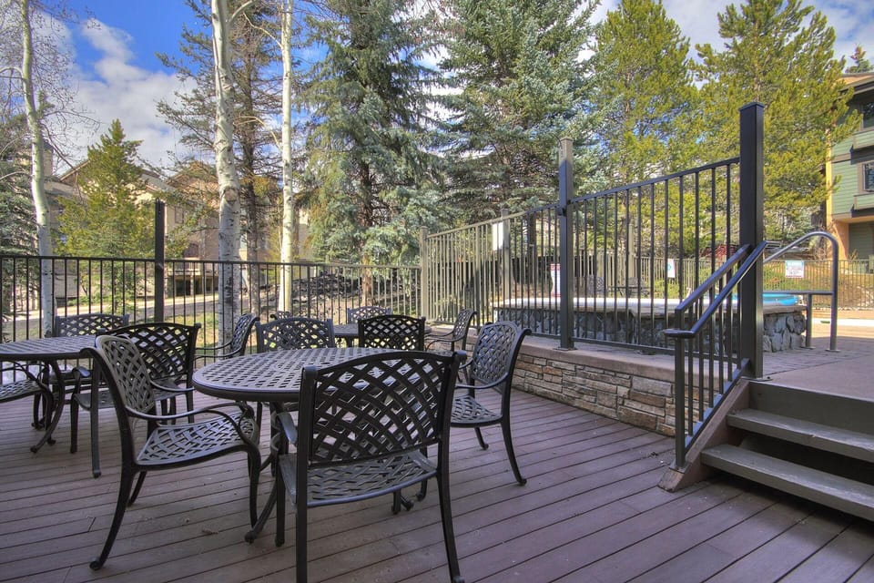 Outdoor patio with black metal tables and chairs on a wooden deck near a hot tub, surrounded by trees and a fence, with buildings visible in the background.