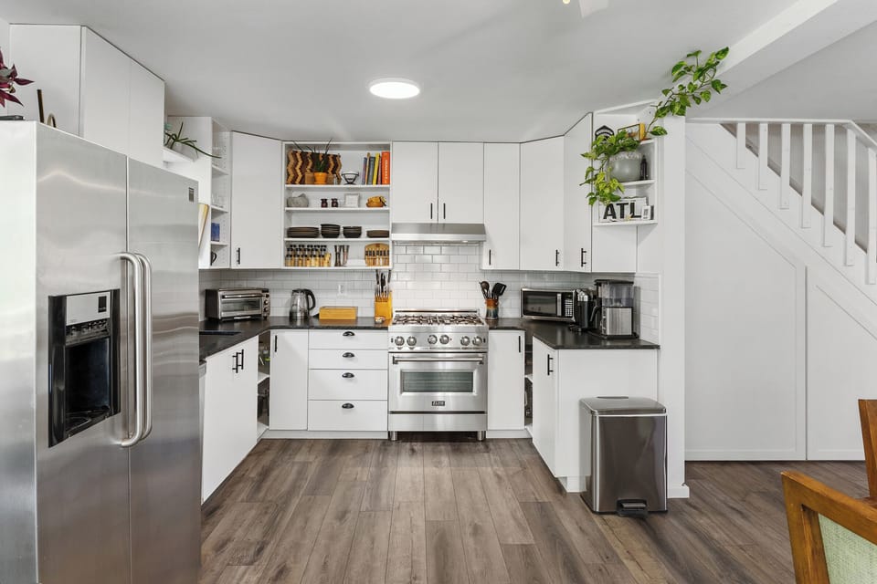Modern kitchen with white cabinets, stainless steel appliances, tiled backsplash, and wooden floors. Plants and decor adorn the shelves and counter. Stairs are visible to the right.
