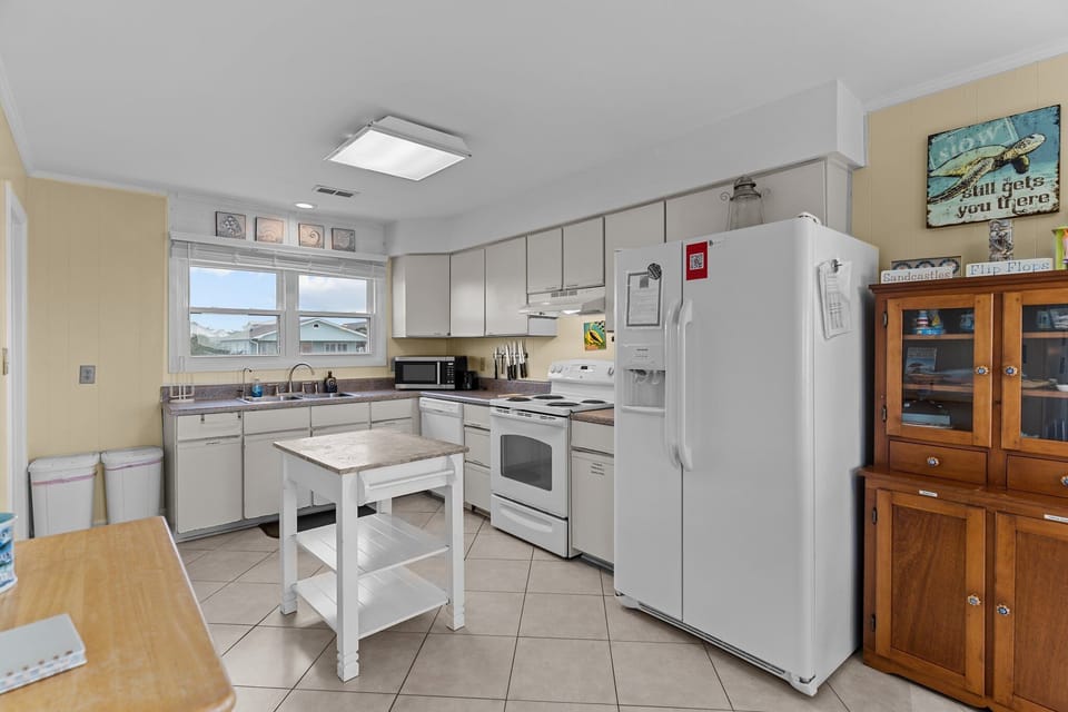 Oak Island, NC kitchen with white appliances, stove, sink, microwave, island cart, tile floors, beach décor, wood cabinet, and bright coastal style.
