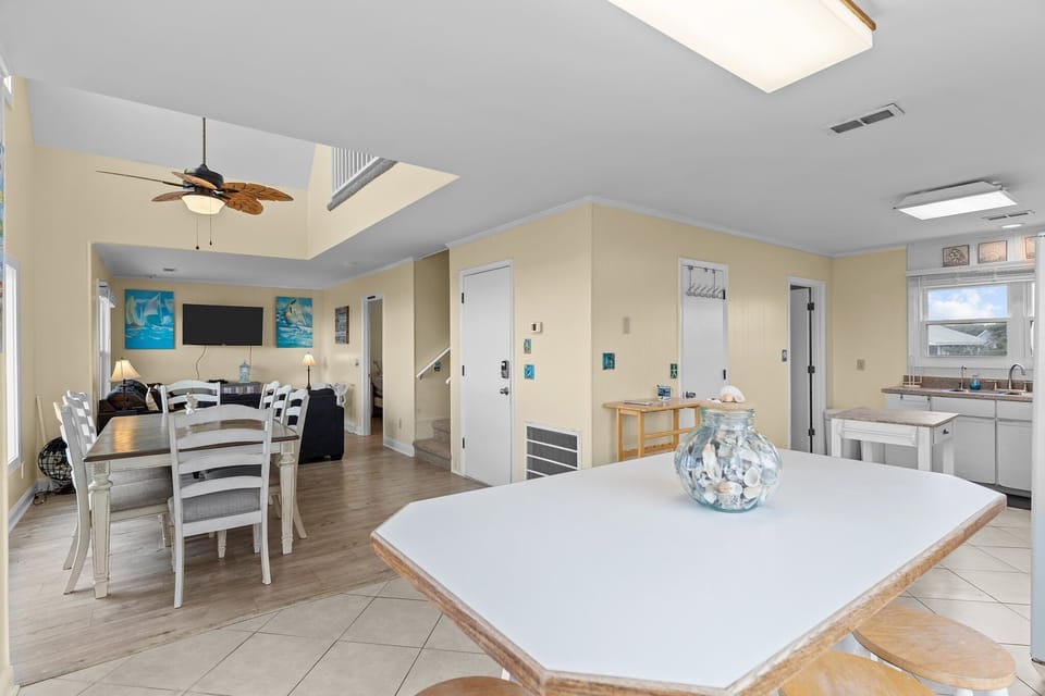 Oak Island, NC dining area with large table, ocean-view window, leaf ceiling fan, breakfast bar, stools, beach décor, tile and wood floors.