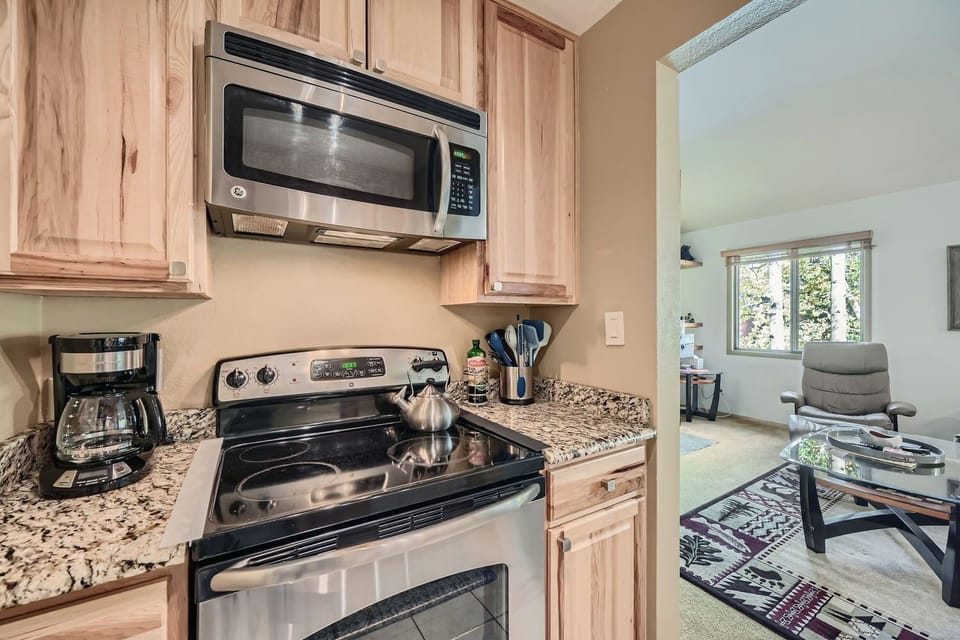 Kitchen with a microwave, electric stove with a kettle, coffee maker on a granite countertop; doorway leading to a living room with a chair and glass coffee table.