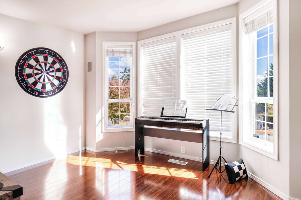 A bright, sunlit corner with a dartboard and a keyboard, perfect for entertainment or practicing music. The room is filled with natural light from the large windows, providing a pleasant environment for relaxation and hobbies.