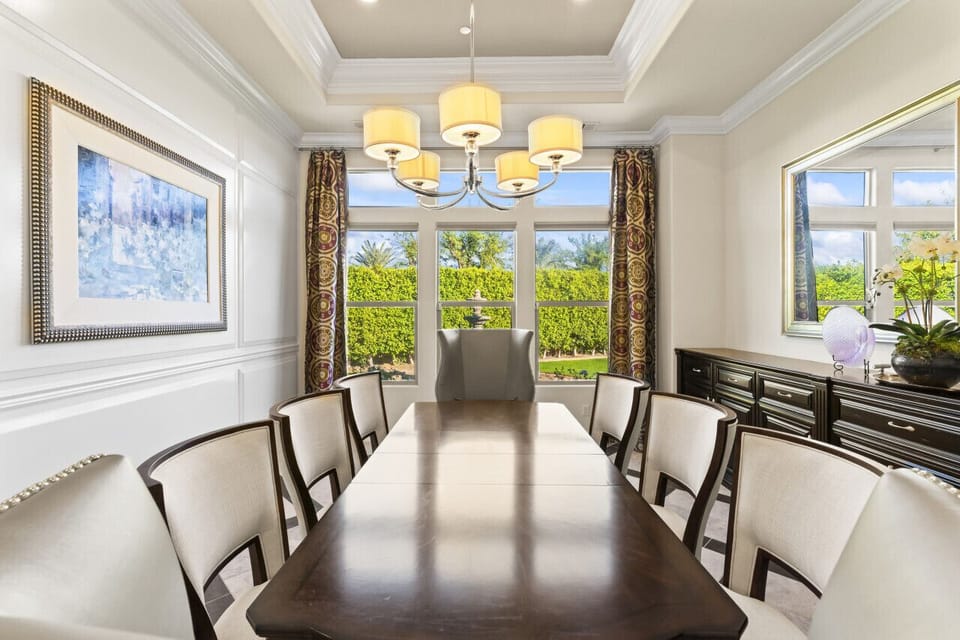 Formal dining room w/ long table, chandelier & large windows framing greenery