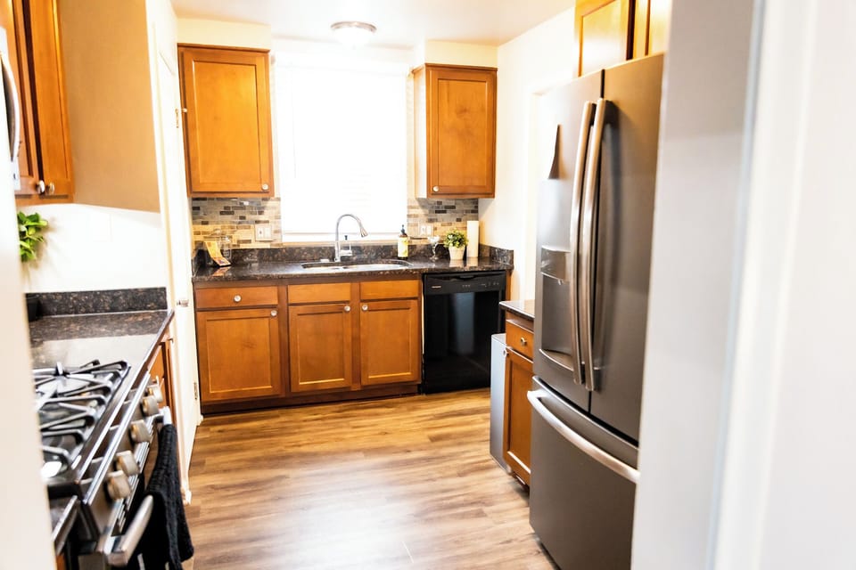 Warm and welcoming kitchen featuring rich wood cabinetry, sleek black granite countertops, and modern stainless-steel appliances. The space is accented by a stylish mosaic tile backsplash and natural light, creating a perfect blend of function/charm.
