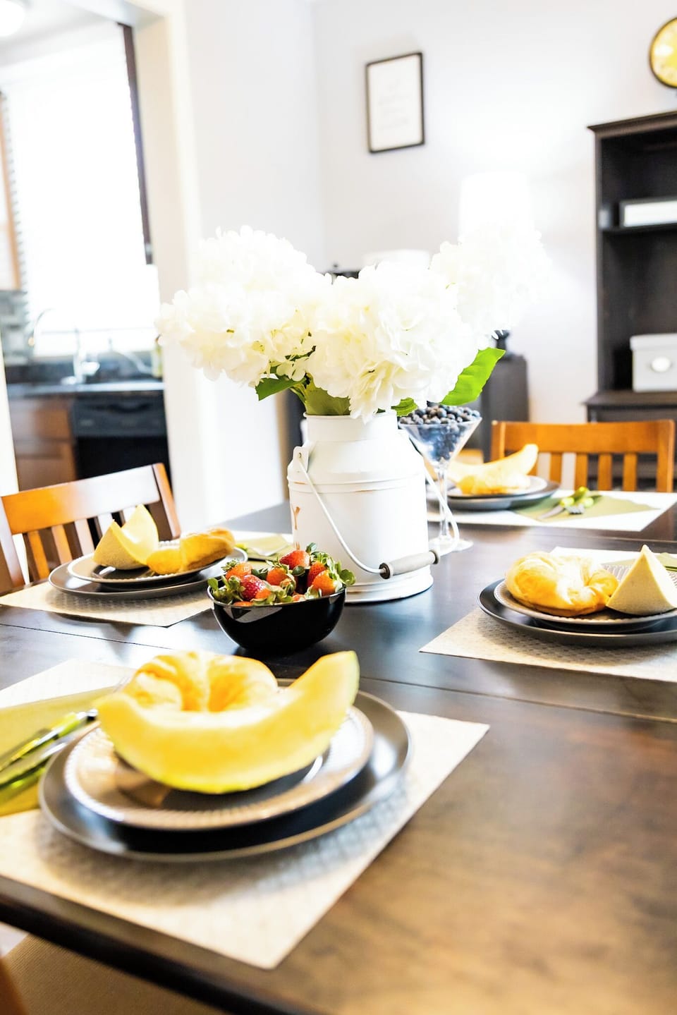 This bright dining room features a dark wood table set with fresh fruit, croissants, and neatly folded napkins. A centerpiece of white hydrangeas in a farmhouse pitcher adds charm, while natural light enhances the cozy yet elegant atmosphere.
