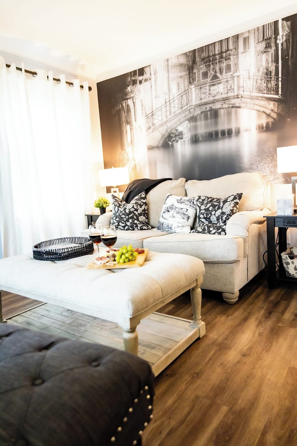 Bright and inviting living room featuring a cozy cream sofa with patterned pillows, a tufted ottoman serving as a coffee table, and a striking black-and-white Venice mural. Perfectly styled with wine, snacks, and soft lighting for a relaxing ambiance