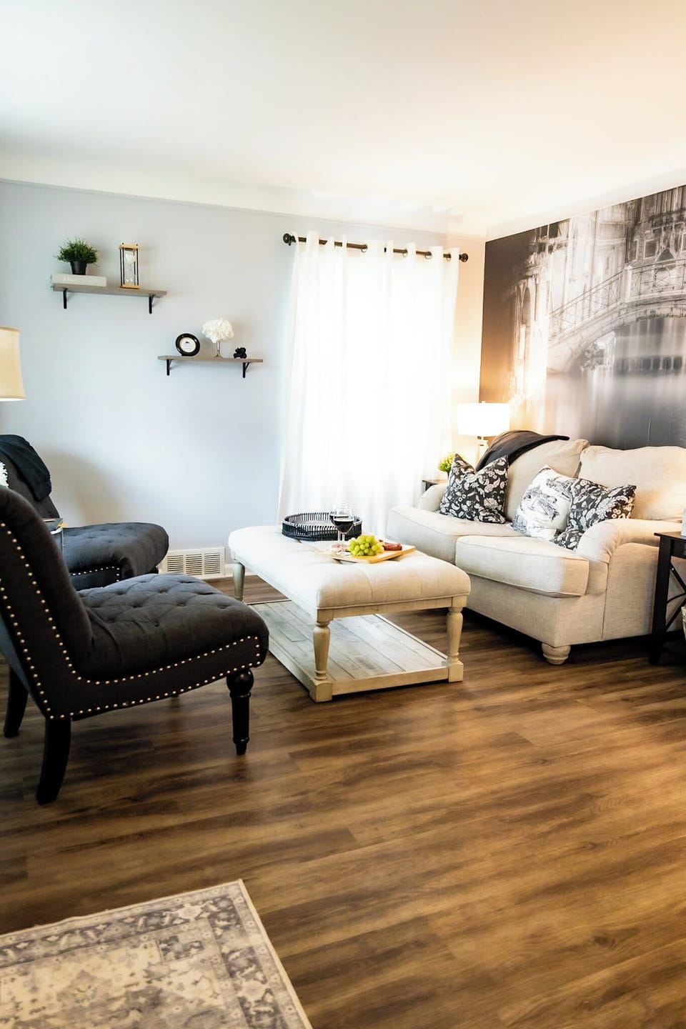 Charming living room with a cozy beige sofa, patterned pillows, and black accent chairs. A large ottoman doubles as a coffee table with snacks and wine, while sheer curtains and wall art add a touch of elegance. Warm wood floors complete tLiving Room