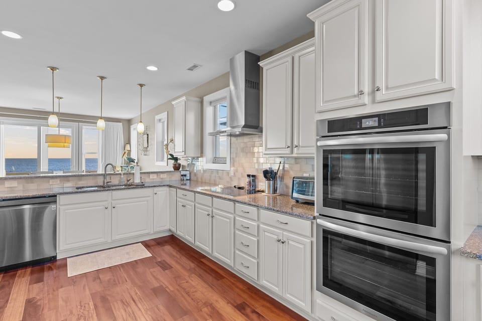 Bright coastal kitchen with white cabinets, double wall ovens, granite counters, stainless appliances, pendant lights, hardwood floors, and ocean views beyond the sink.