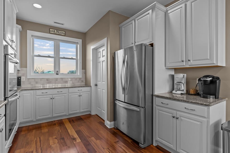 Bright coastal kitchen with white cabinets, double wall ovens, granite counters, stainless appliances, pendant lights, hardwood floors, and ocean views beyond the sink.