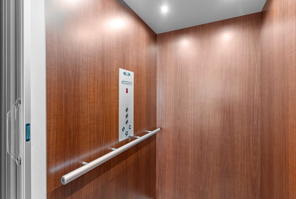 Private residential elevator with warm wood-paneled walls, recessed lighting, metal handrail, and control panel for easy access between floors.