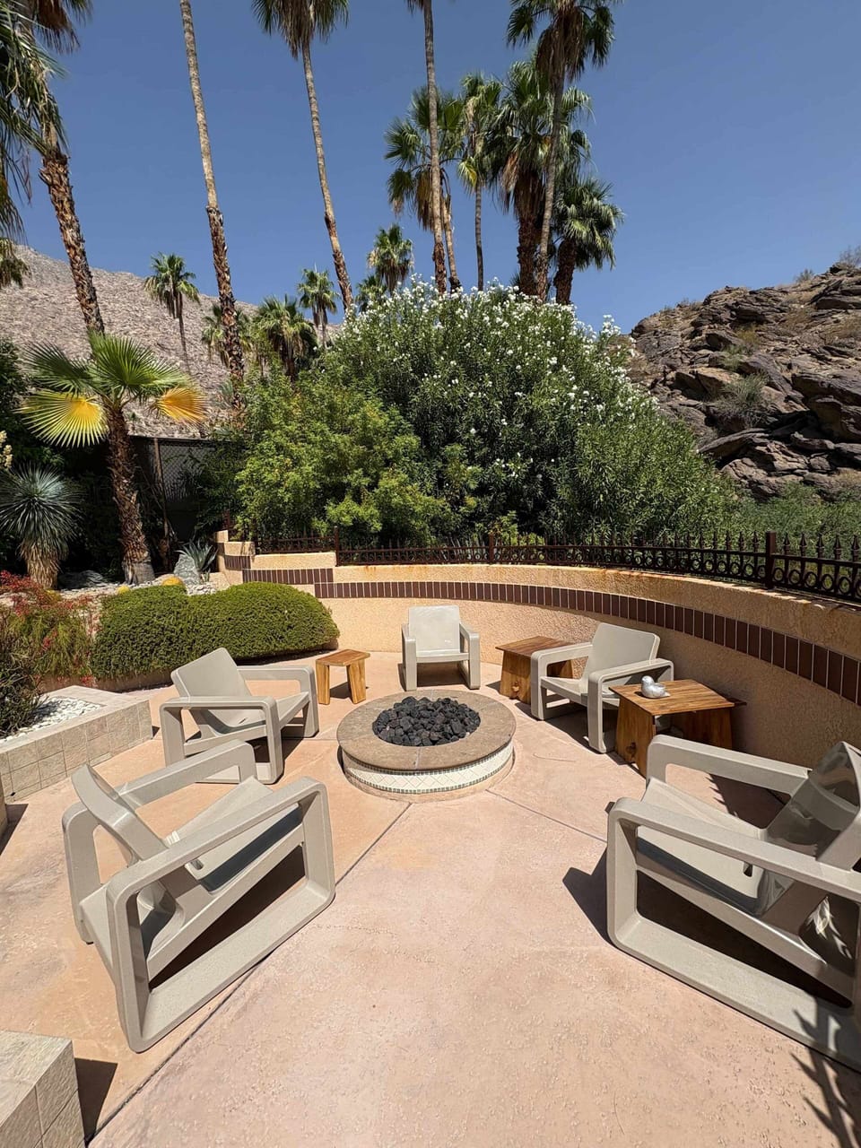 Sunny outdoor fire pit patio with six lounge chairs, palm trees, and a dramatic boulder backdrop — desert evenings done right