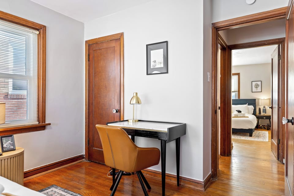 Bedroom view with a compact desk and chair beside the window, hardwood flooring, and a sightline through the hallway to the second bedroom.
