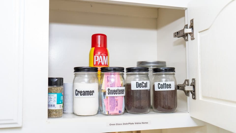 Conveniently stocked kitchen cabinet with essentials for your morning routine, including labeled jars of coffee, decaf, creamer, and sweetener. Also features cooking spray and seasonings making it easy to prepare meals and enjoy fresh coffee anytime.